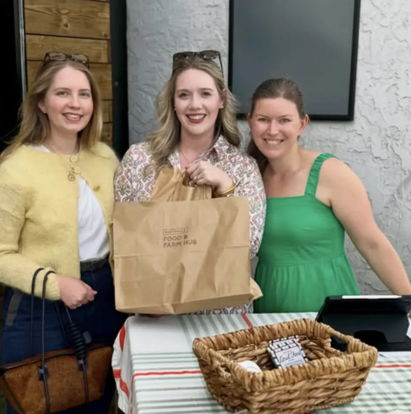 Three women smiling outside while holding a Huntsville Food & Farm Hub bag.